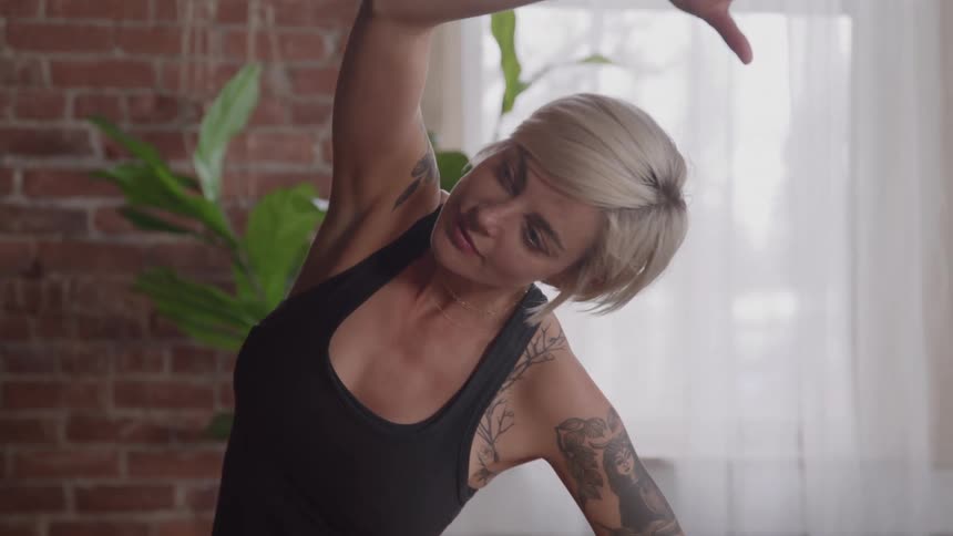 Shorthaired woman with tattoos does a side stretch indoors