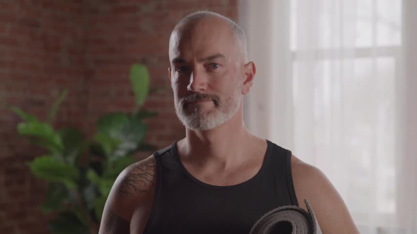 A man with a shaved head and gray beard wears a black tank top and holds a rolled up yoga mat under his left arm turning his head to look directly at the camera