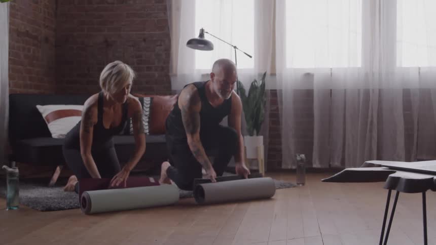 A wide shot shows a man and woman setting up for a workout in a loft apartment with exposed brick walls they are each carrying a yoga mat and a water bottle and they place their gear on the floor