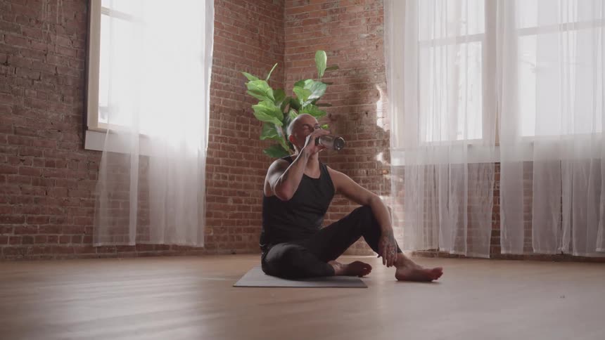 Man sitting on a yoga mat drinking water in a bright room