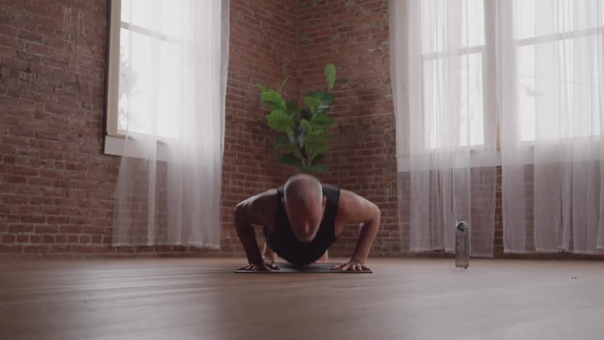 A man with a shaved head and tattoos on his arms is in a room with a brick wall and two large windows doing a pushup on a yoga mat with a water bottle on the floor nearby
