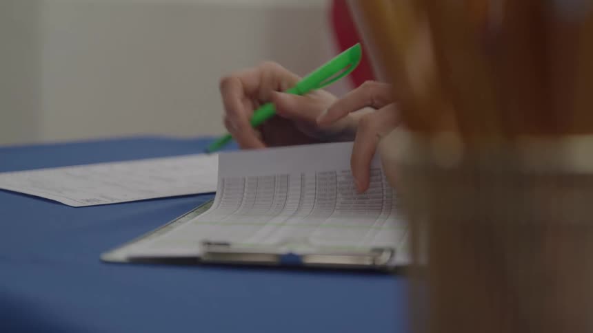 A person wearing a striped shirt and holding a green highlighter writes on a clipboard with a checklist on top of a blue table with an american flag visible behind them