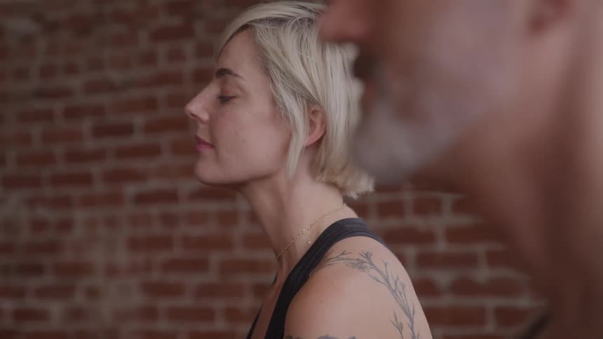 A young woman with a tattoo on her back and a middleaged man sit indoors at midday doing yoga