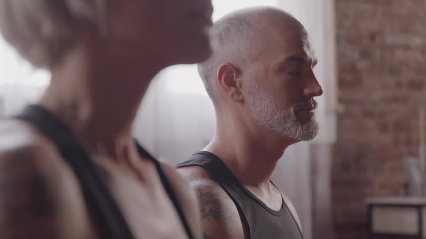 A male and a female are seen from the chest up sitting in a yoga pose with their eyes closed they are practicing meditation or mindfulness in what looks like an urban loft with brick walls and large windows