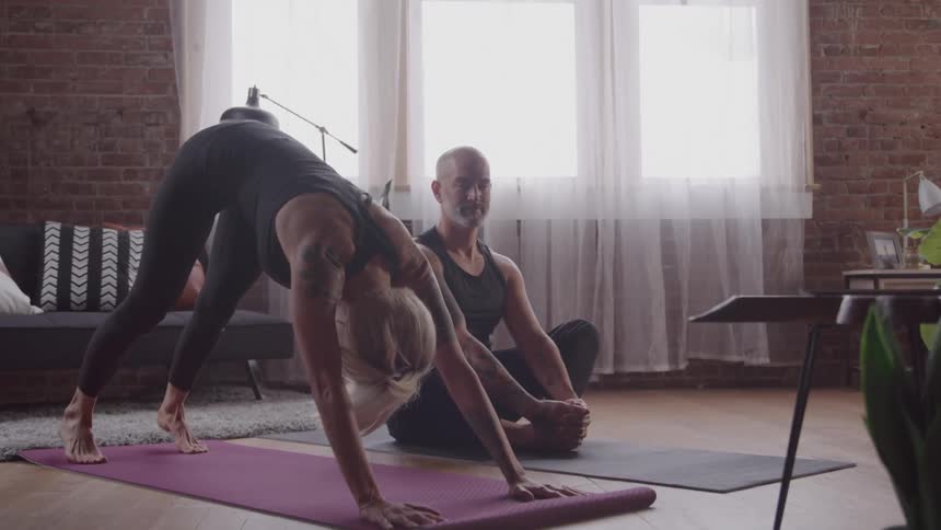 Two people practice yoga in a bright room with large windows