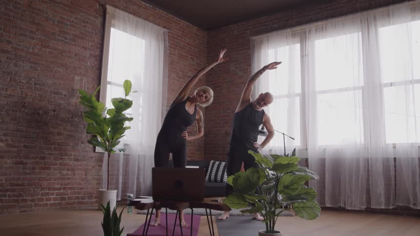 An elderly couple wearing similar spandex exercise clothes do stretches in a corner of a large room with an opened laptop in front of them
