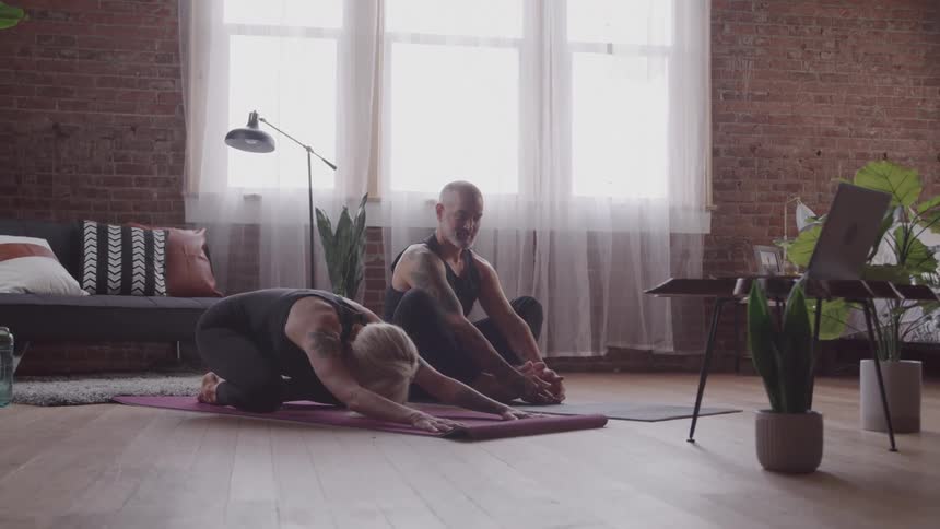A bald man and a woman with gray hair are doing yoga together in their living room she is in a childs pose with her head down while he sits with his feet pressed together watching a yoga session on his laptop