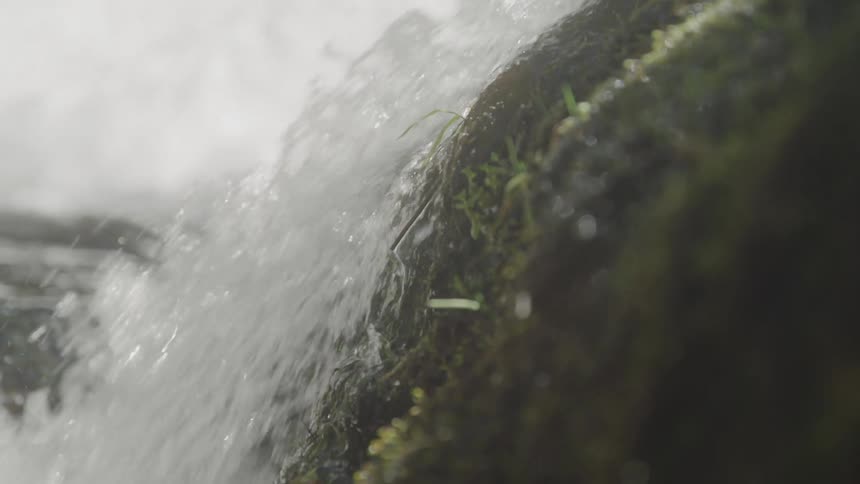 A closeup panning shot follows whitewater as it rushes down the side of a mossy rock in a stream or waterfall