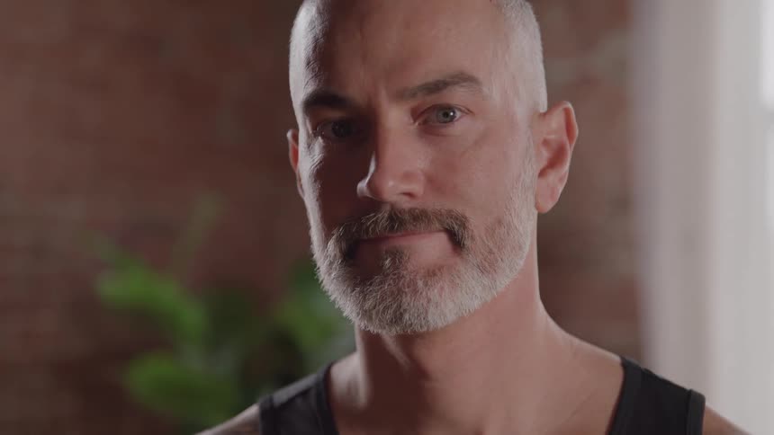 A man with a shaved head and graying beard is shown in a closeup side profile he then turns his head to face the camera looking directly at the viewer with a calm expression