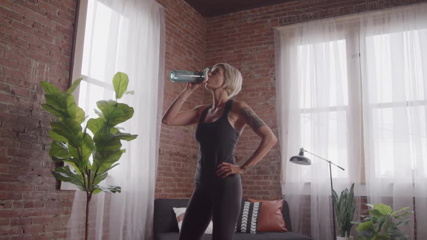 A woman with blonde hair and a tattoo on her left arm stands in a room with exposed brick walls and large windows drinking from a translucent blue water bottle