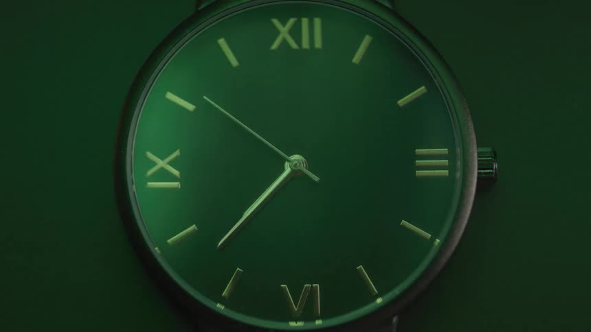 A watch with a round green face and yellow gold hands and roman numerals with the second hand visibly sweeping around the face