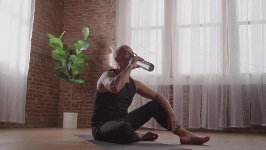Man sitting on a yoga mat drinking water from a bottle in a bright room