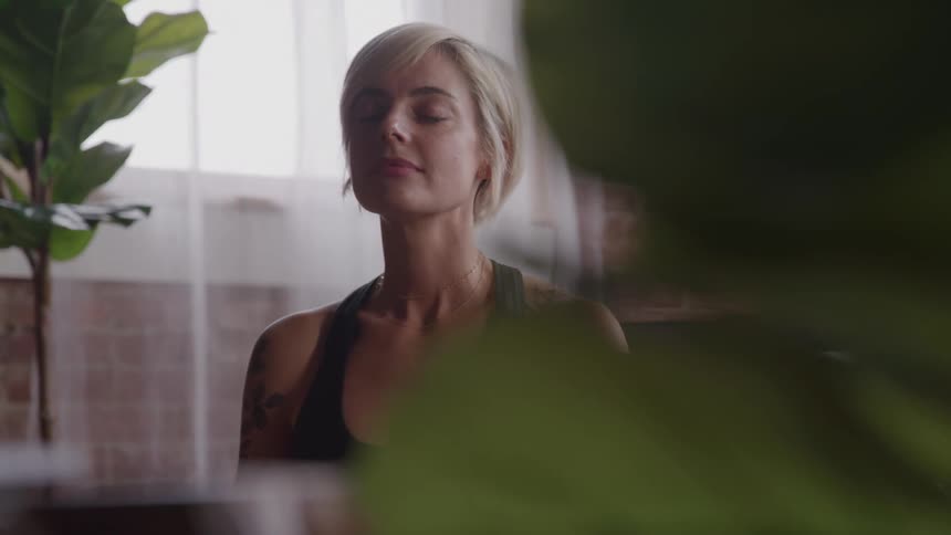 A shorthaired blonde woman is sitting with her eyes closed in what appears to be a meditative state she is wearing a dark tank top and is partially obscured by a large green plant in the foreground