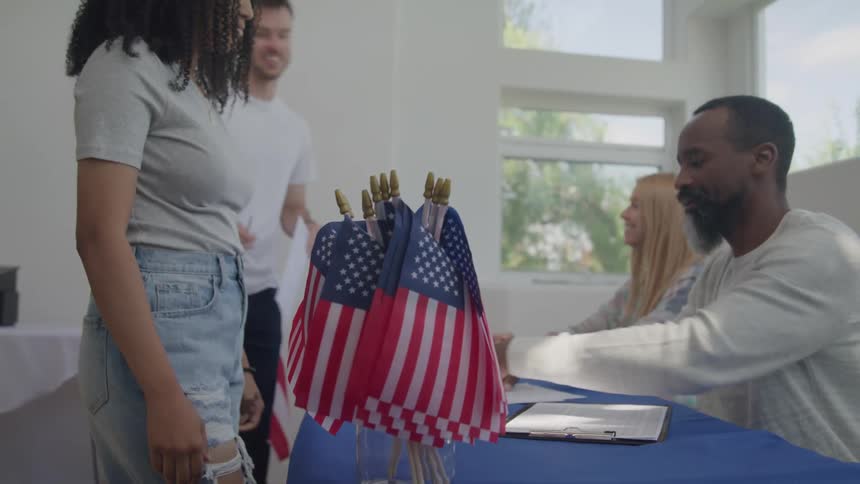 A shot of a line of people in a room decorated with american flags and people waiting to get a paper to vote