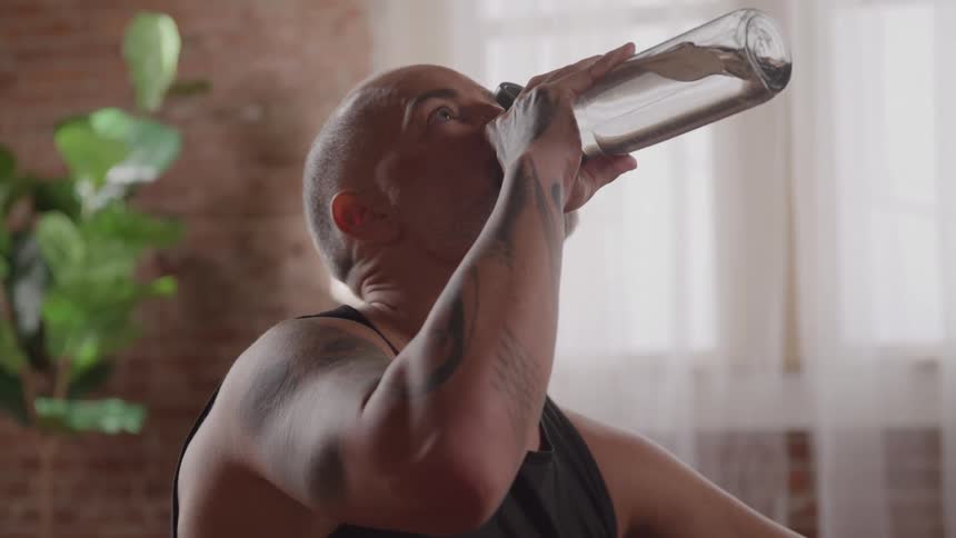 A middleaged bald man with a beard is shown from the chest up he takes a long drink from a reusable water bottle then lowers it and looks forward appearing relaxed after a workout