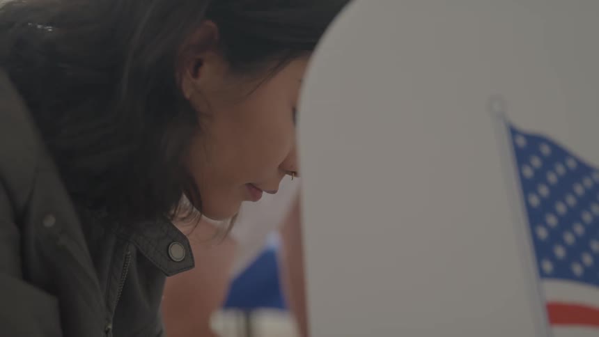 A person with long dark hair is shown in closeup standing in a voting booth they are focused on filling out their ballot with the corner of an american flag visible in the background