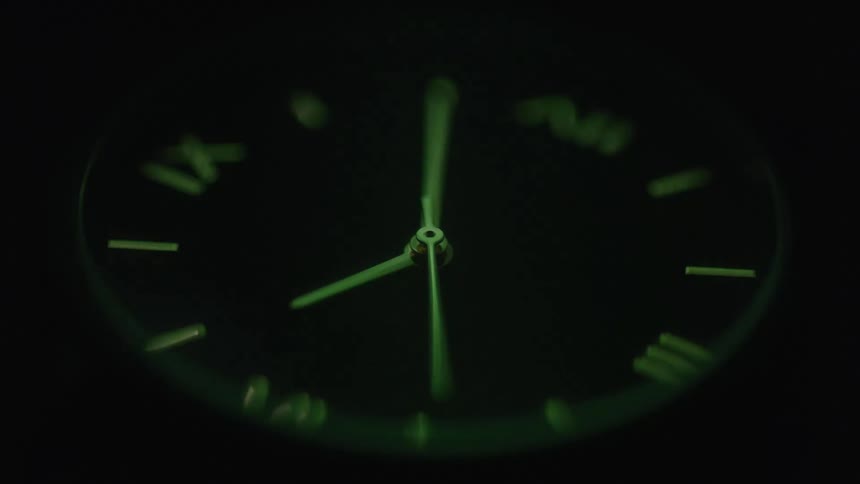 A macro shot of an analog clock face shows the second hand sweeping across the dial the numbers and hands are luminous glowing in the dark