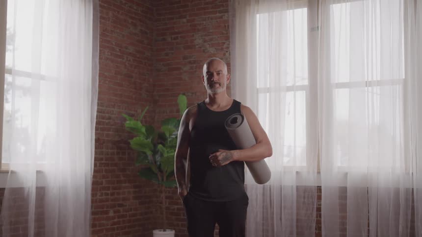 A middle aged man stands in a living room during daytime while holding a yoga mat