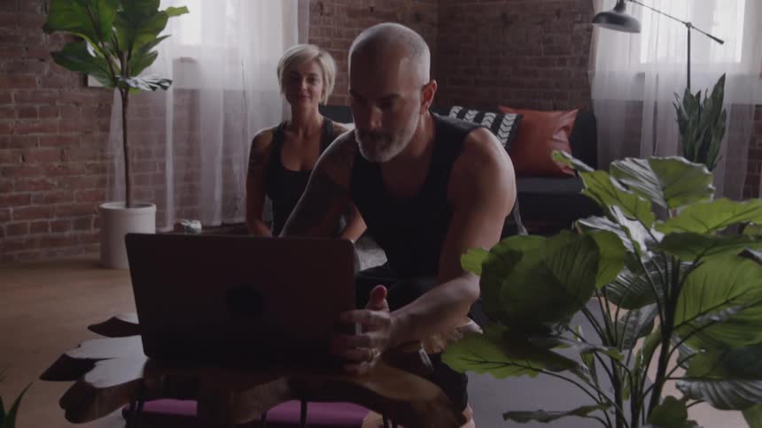 A male and female yoga practitioner are meditating in a loft apartment with brick walls and large windows the woman is seated in a yoga pose with her eyes closed while the man also with his eyes closed is positioned slightly behind her a laptop is ope