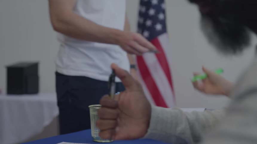 Two poll workers sitting side by side checking peoples ids before handing out ballot papers for them to cast their votes with an american flag on the wall inside the polling station