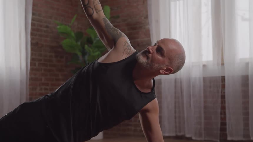 A bald man with tattoos reaches upwards to perform a yoga pose