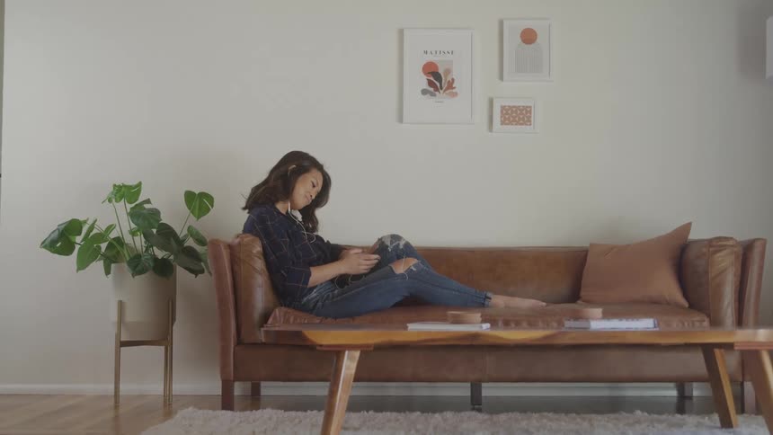 A young woman listens to music with earphones and uses a mobile phone while sitting on a couch in a living room during daytime