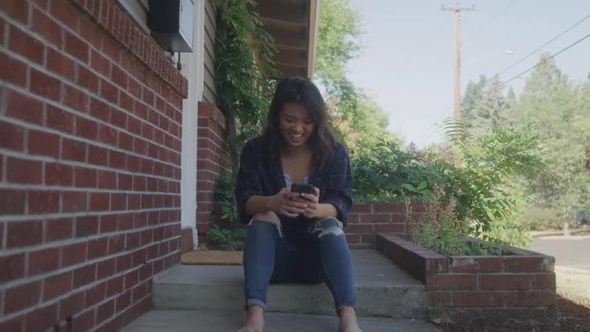 A wide shot shows a young woman sitting on the front steps of a brick building she is looking down at her cell phone and has a slight smile on her face