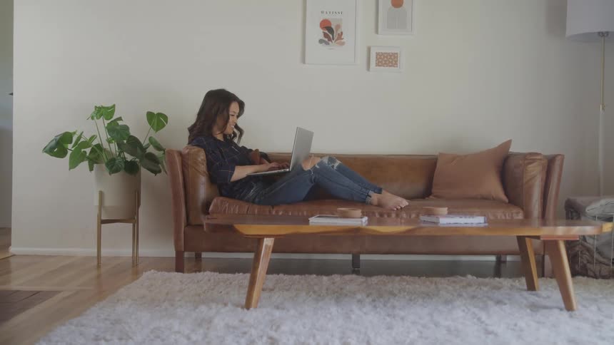 A woman with dark hair sits on a brown leather sofa typing on a laptop