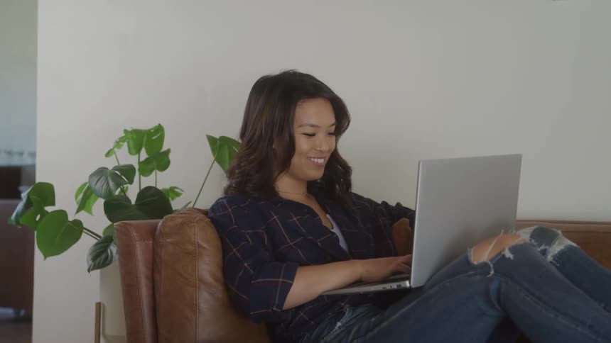 A person with dark mediumlength hair sits comfortably on a brown leather couch they are smiling while looking at a laptop that is resting on their lap