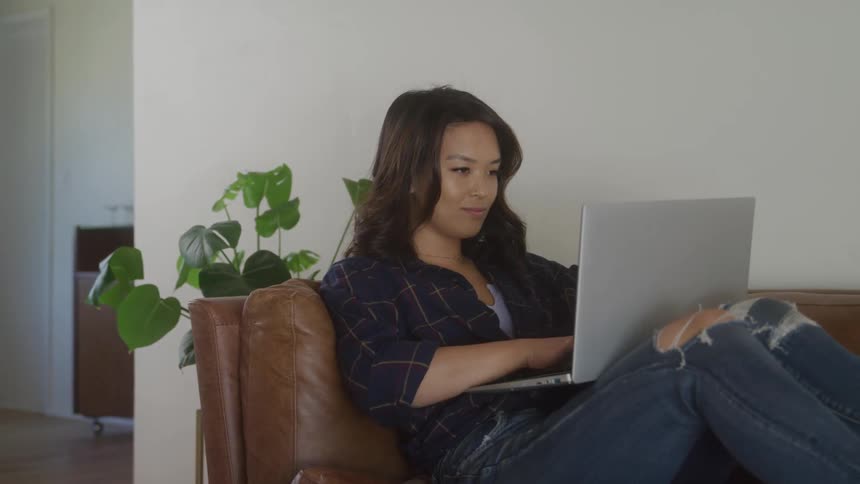 A young woman sits on her couch with a laptop on her lap typing with a flower pot behind her