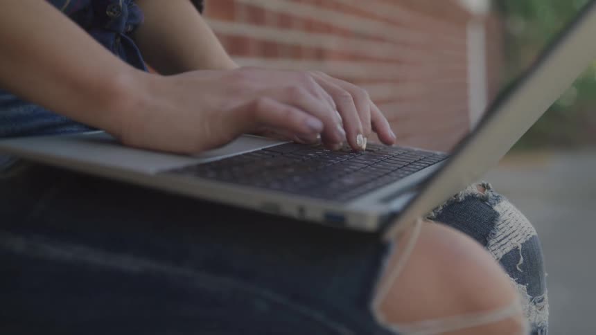 A person wearing torn blue jeans types on a laptop resting on their lap with their fingers moving across the keyboard