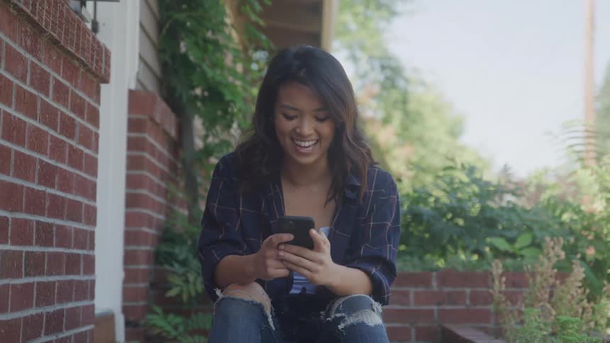 A woman with dark hair sits on the brick steps of a house smiling as she looks down and uses her cell phone