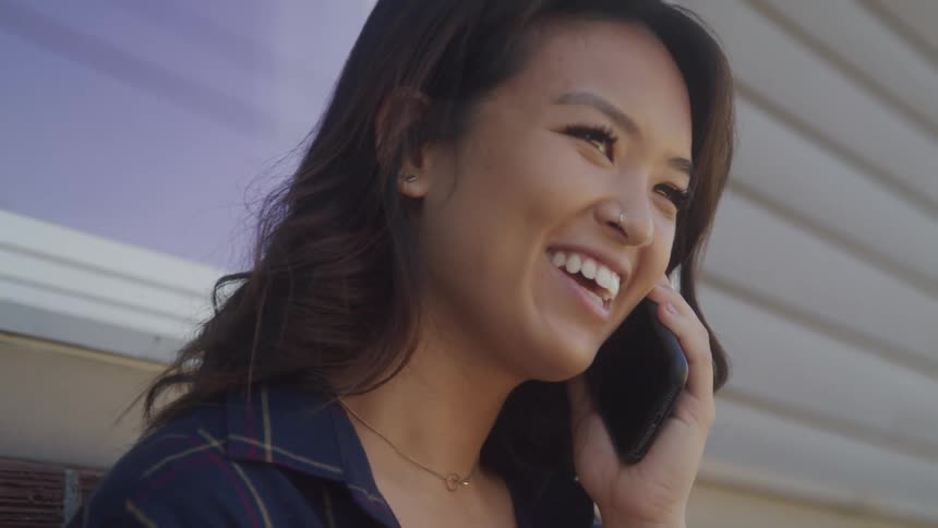 A young woman is shown in a closeup chestup shot while talking on her cell phone she has a large smile on her face and is leaning against a brick wall