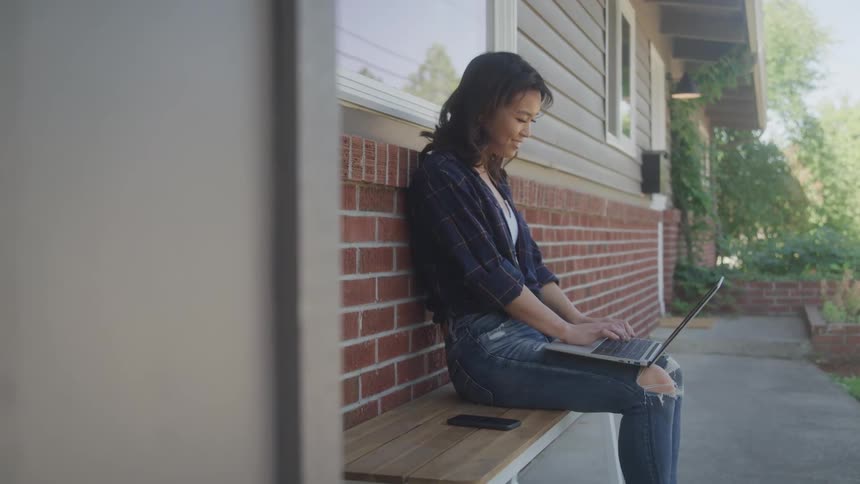 A woman with long dark hair sits on a wooden bench on a front porch smiling as she types on a laptop while a cell phone rests on the bench beside her