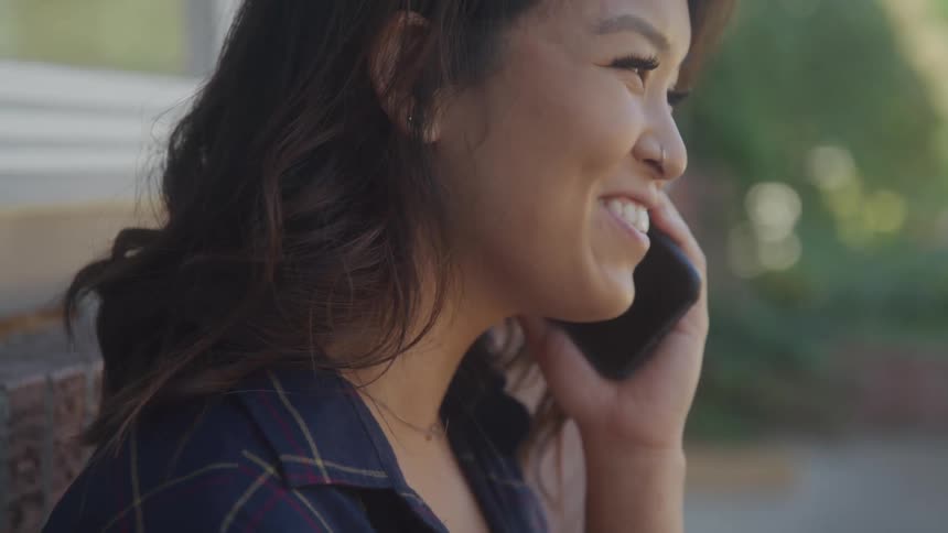 Smiling woman talks on the phone leaning against an outside wall