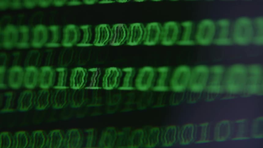 A closeup shot of a computer screen displays scrolling lines of green binary code on a black background representing data or hacking