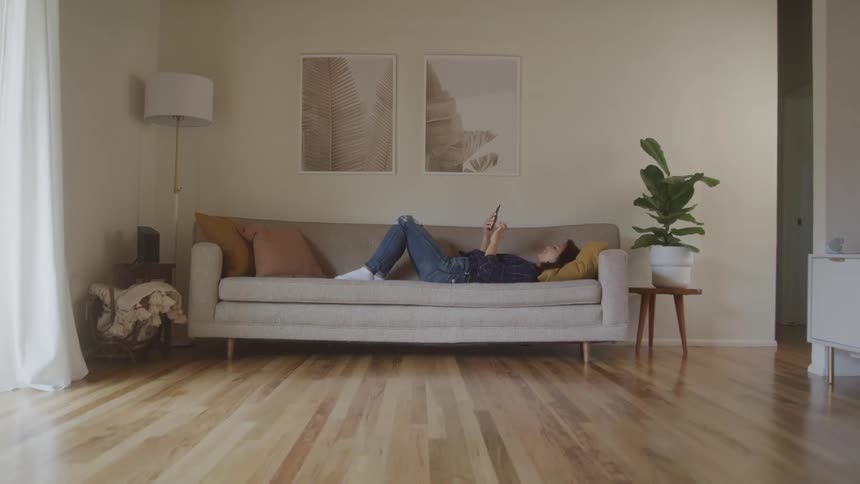 A young person with a yellow beanie and blue jeans is lying on a lightcolored couch engrossed in a cell phone the room is brightly lit with hardwood floors and minimalist decor