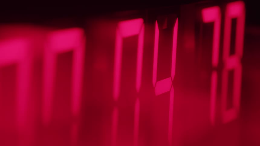 In an angled closeup shot with a shallow depth of field the red segmented numbers of a digital clock rapidly count down to zero