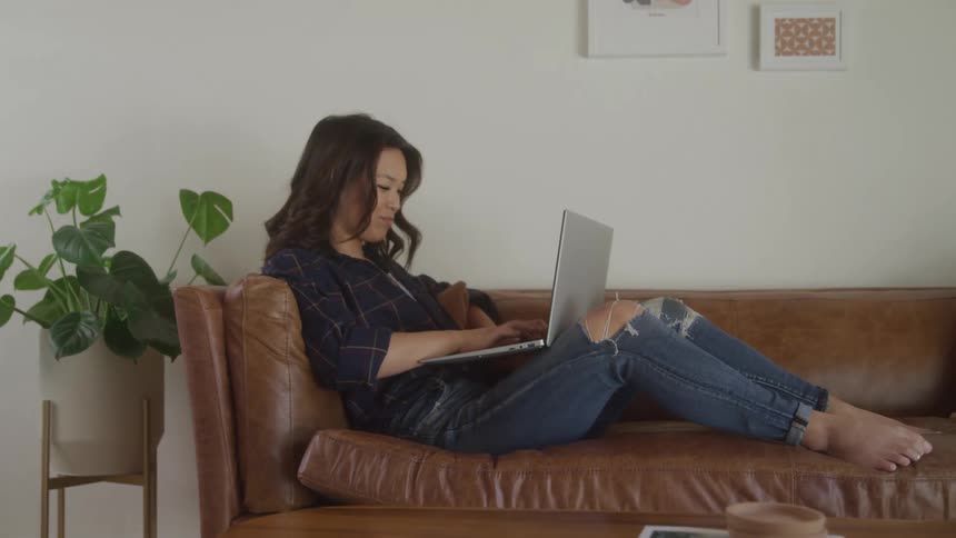 A young woman uses a laptop computer while sitting on a couch in a living room during daytime
