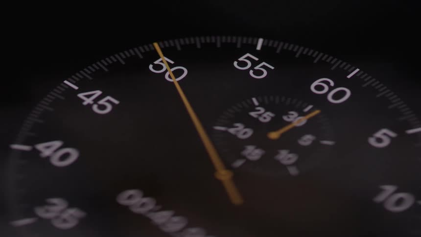 In an extreme closeup the yellow second hand of a black analog stopwatch sweeps smoothly across the numbered dial precisely measuring the passage of time