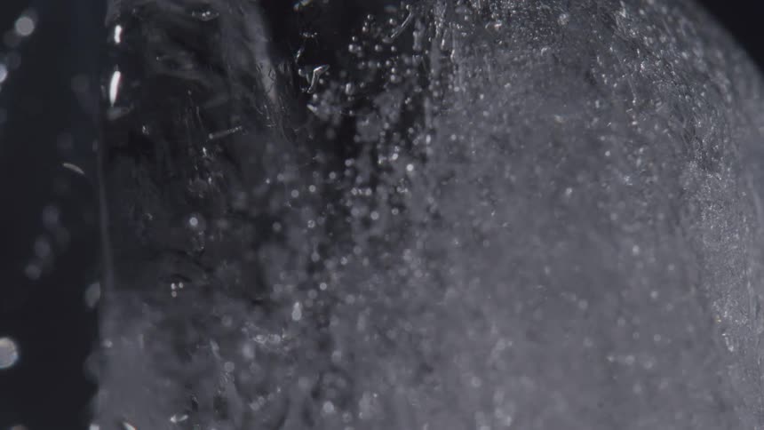 A macro video shows bubbles fizzing on the textured surface of a piece of ice before it begins to visibly melt and trickle with water