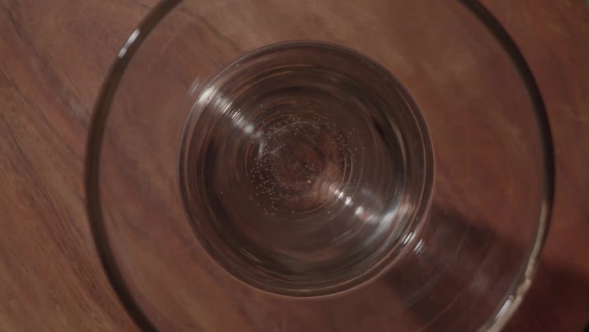 A static topdown view shows tiny bubbles rising through a clear sparkling beverage in a glass resting on a wooden surface
