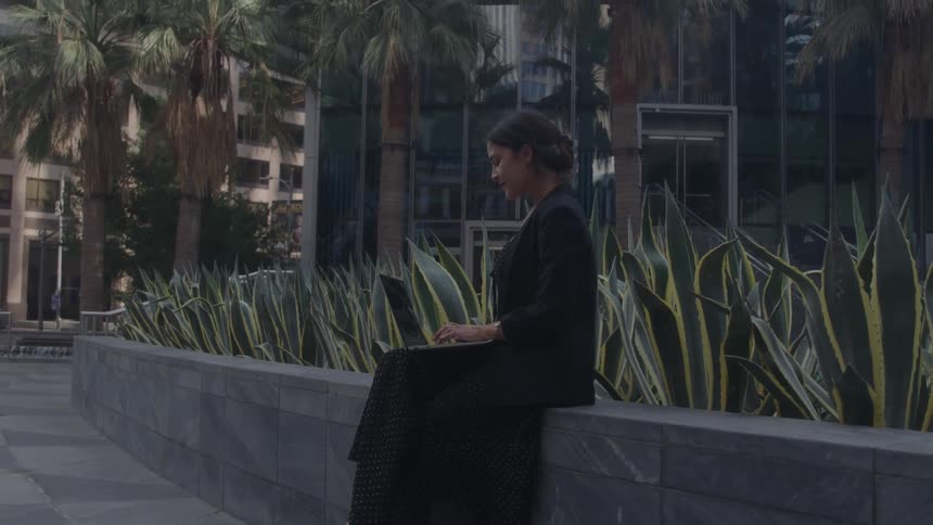 A young woman sits on a low wall in an urban setting looking at her laptop behind her is a modern glass building and a row of palm trees