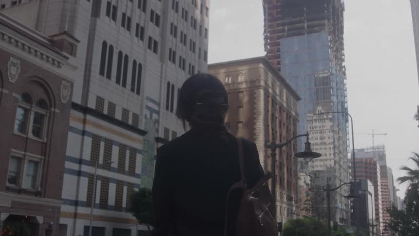 A person with a dark jacket stands on a city sidewalk lined with tall buildings
