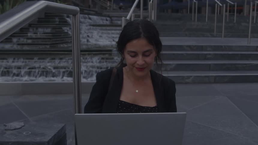 Woman working on a laptop seated in front of a fountain with urban stairs in the background