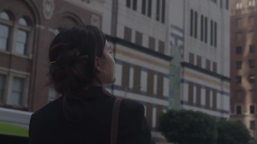 A woman with dark hair and a dark jacket standing on a city street and looking up at tall buildings