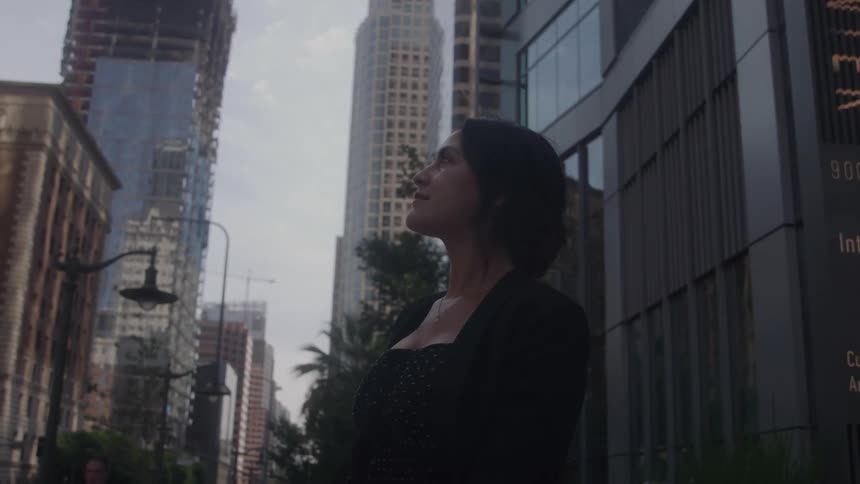A woman with short dark hair stands on a city sidewalk and looks up at the tall buildings around her