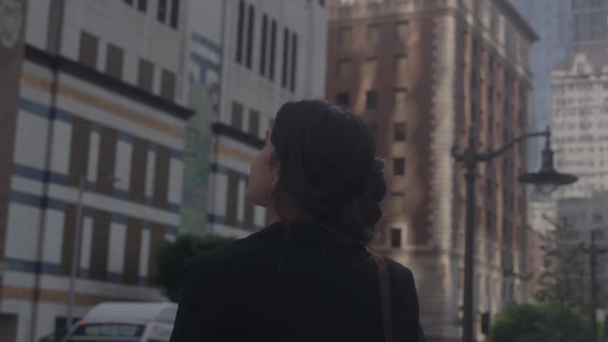 A woman with a dark jacket and a bun in her hair stands on a city street and looks up at a tall light brown building with many windows and a single streetlamp on the sidewalk