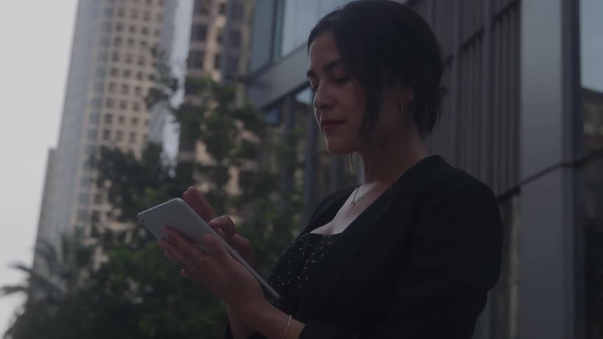A woman with her hair pulled back and wearing a black jacket stands outdoors in a city looking down and scrolling on a tablet she holds in her hands