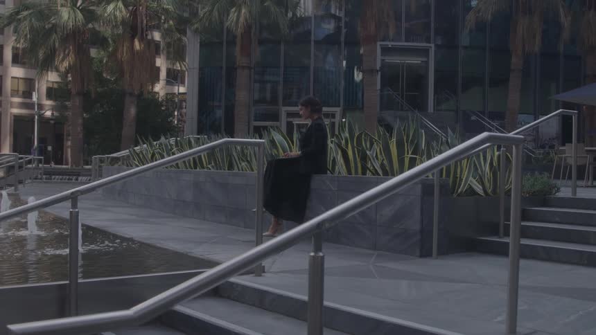 A young woman uses a laptop computer while sitting on a pavement outdoors beside a sidewalk in a metropolitan city during daytime
